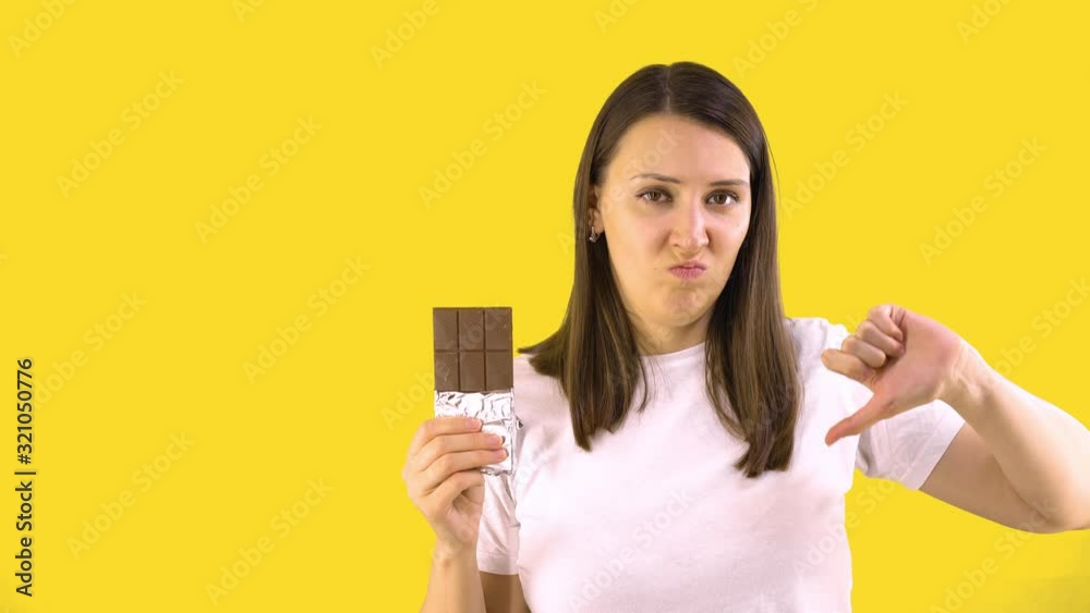 Young Woman With a Chocolate Bar, Shows That it is Harmful. Yellow Background. People sincere emotions lifestyle concept. Young Woman With a Chocolate Bar, Shows That it is Harmful. Yellow Background. People sincere emotions lifestyle concept.