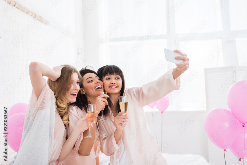 attractive multicultural girls with glasses of champagne taking selfie on smartphone during pajama party attractive multicultural girls with glasses of champagne taking selfie on smartphone during pajama party