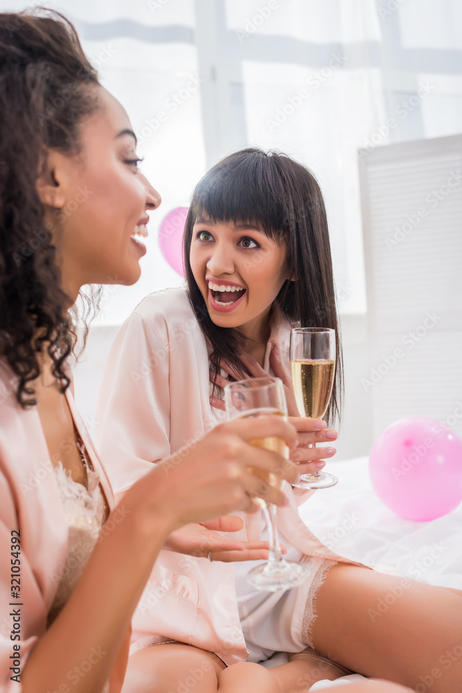 surprised multicultural girlfriends with glasses of champagne talking on pajama party surprised multicultural girlfriends with glasses of champagne talking on pajama party