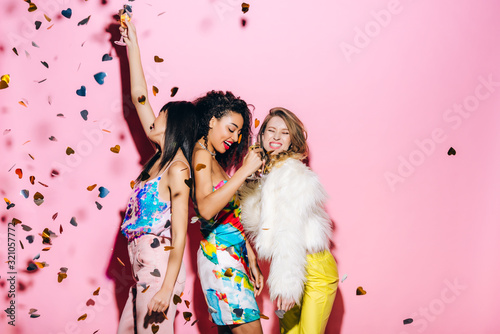 Excited multicultural girls dancing with glasses of champagne on pink with confetti photo