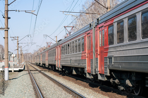 Russian electric train in red, orange, gray. Railway, poles with wires. Spring day