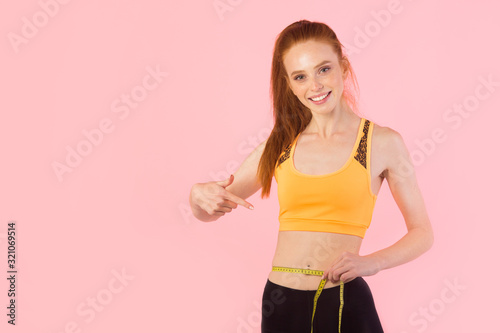 beautiful young slim woman with red hair in sportswear on a pink background with a measuring tape