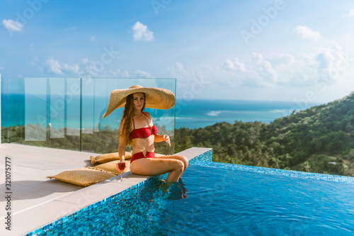 A beautiful girl in a red bathing suit is relaxing in a luxury villa by the pool with wine and in a hat taking cover from the sun. View of palm trees and the sea. Thailand, Koh Samui