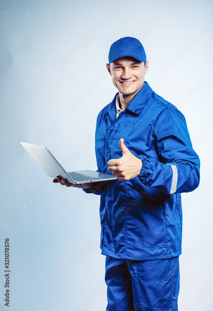 Portrait of young worker man wearing blue uniform. Holding a laptop in his hand and showing thumb up. Movement cool. Isolated on grey background with copy space. Business concept. Portrait of young worker man wearing blue uniform. Holding a laptop in his hand and showing thumb up. Movement cool. Isolated on grey background with copy space. Business concept.