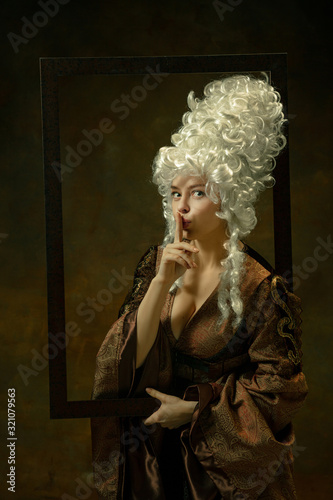 Whispering secret. Portrait of medieval young woman in vintage clothing with wooden frame on dark background. Female model as a duchess, royal person. Concept of comparison of eras, fashion, beauty.