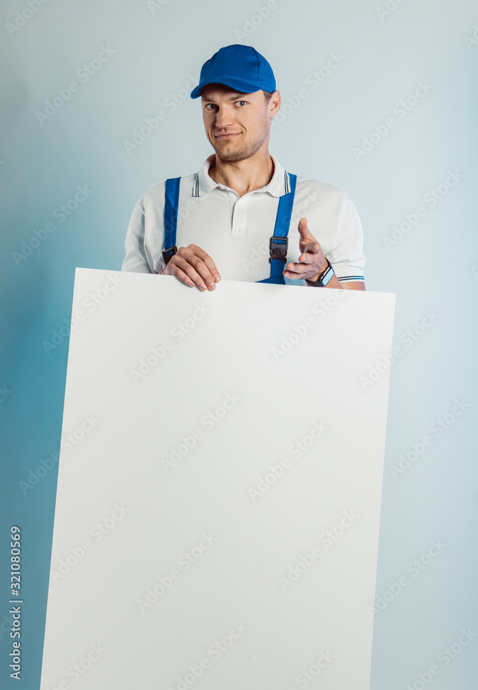 Mockup image of young smiling worker man wearing blue uniform. Holding white empty banner in his hand and pointing finger at you looking at camera. Isolated on grey background with copy space. Busines Mockup image of young smiling worker man wearing blue uniform. Holding white empty banner in his hand and pointing finger at you looking at camera. Isolated on grey background with copy space. Busines