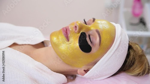 Hand of professional cosmetologist applying gold mask on client's face.