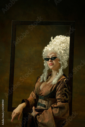 Trying on eyewear. Portrait of medieval young woman in vintage clothing with wooden frame on dark background. Female model as a duchess, royal person. Concept of comparison of eras, fashion, beauty.