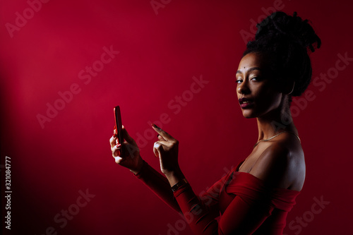 Studio portrait of a woman with dreadlocks wearing a sparkling dress in a studio portrait with red background. Texting on her mobile phone