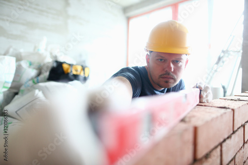 Portrait of skilled professional bricklayer using special equipment and tools to measure balance and height of brick wall. Prudent builder wearing hard gloves to keep hands unharmed. Building concept photo