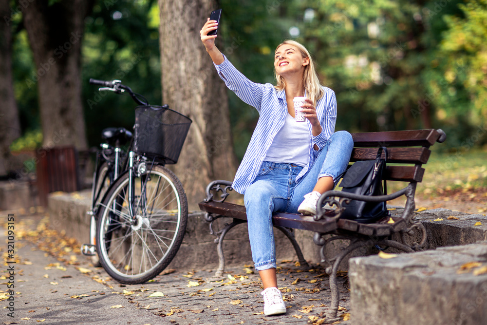 Blonde young girl with coffee to on hand taking selfie by mobile phone in part with bicycle on background Blonde young girl with coffee to on hand taking selfie by mobile phone in part with bicycle on background