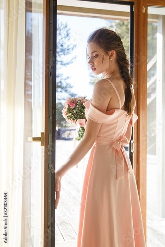 Portrait of a young bride girl in a gently flowing pink dress with a bouquet in her hands stands at the door to the street on a sunny day.