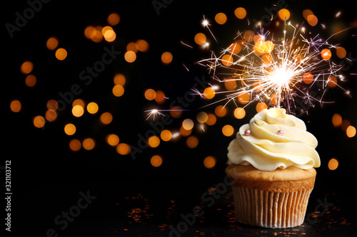Delicious birthday cupcake with sparkler on black table against blurred lights. Space for text