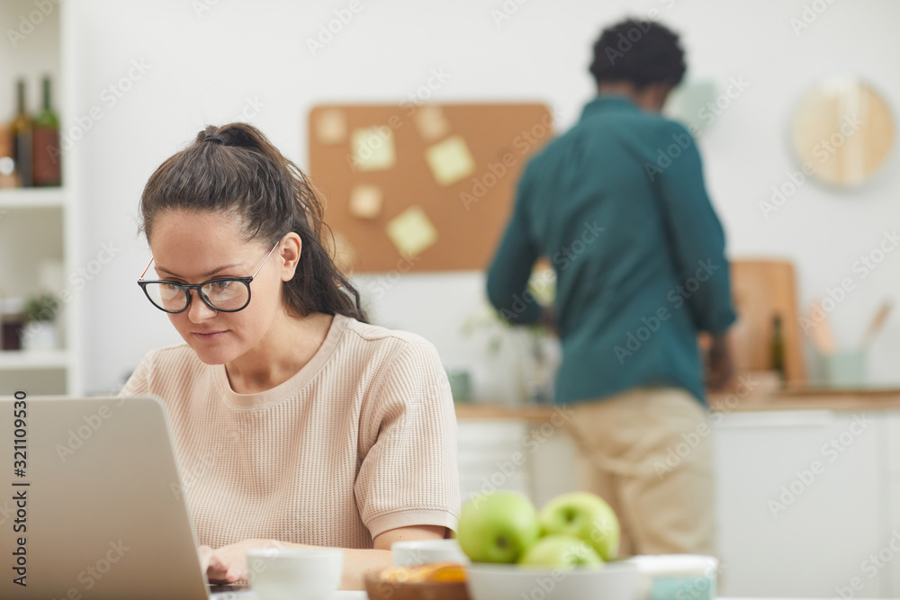 Young woman in eyeglasses typing on laptop while sitting at the table in the kitchen with young man in the background