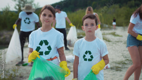 Young kids volunteers collecting litter into plastic bags with ecologu team. Group portrait of two cute children helping environment fighting with pollution. Helpful young generation.