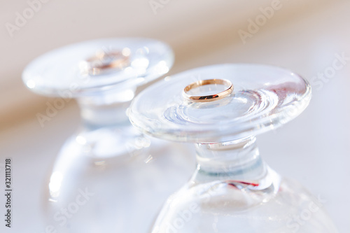 Top view on golden wedding rings lying on top of upside down staying wine glasses. Sun shines through transparent glass.