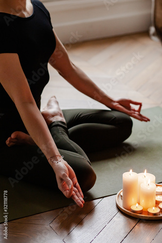 Close-up. Woman in yoga lotus pose meditating in a dark room with candle light. Atmosphere of relax and zen. Exercise to reach clarity of mind and perfect body. Wooden floor, soft morning light