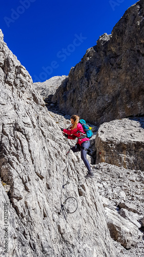 A woman with big hiking backpack climbing on a very steep via ferrata to reach the Grosse Gamswiesenspitze in Lienz Dolomites, Austria. Sharp and barren slopes. Massive Alpine mountains. Solo wanderer
