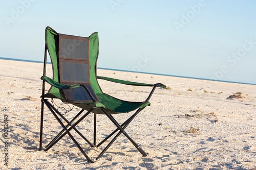 A portable foldable solar panel hangs on a travel chair. Tourist camping on the seashore.