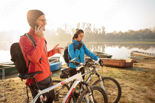 Beautiful young couple riding mountain bikes by the lake in early morning. Man is talking on the phone, woman drinking water. Sun rising through the fog in the background