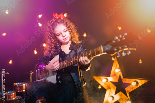 Portrait of a Beautiful rock girl with curly hair wearing leather jacket, boots playing the electric guitar behind a red tank in recording studio.