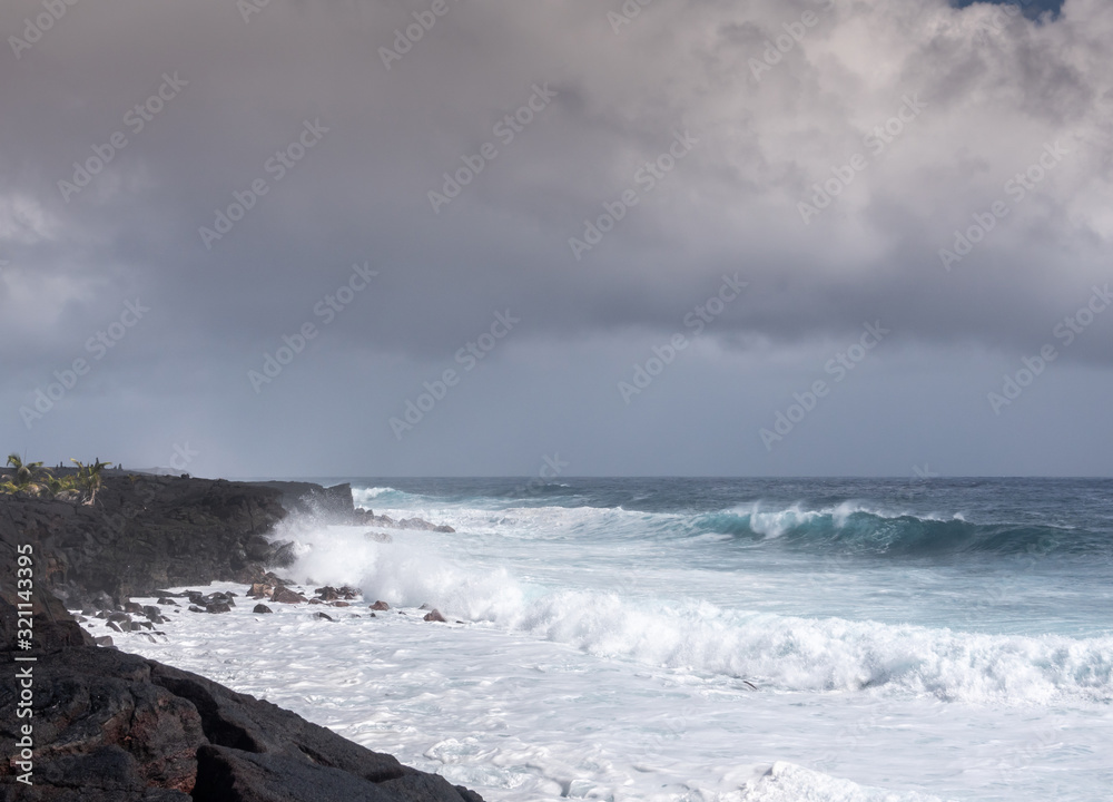 Kaimu Beach, Hawaii, USA. - January 14, 2020: Azure wave crashes with white surf on black lava coastline under heavy rain brown cloudscape. 