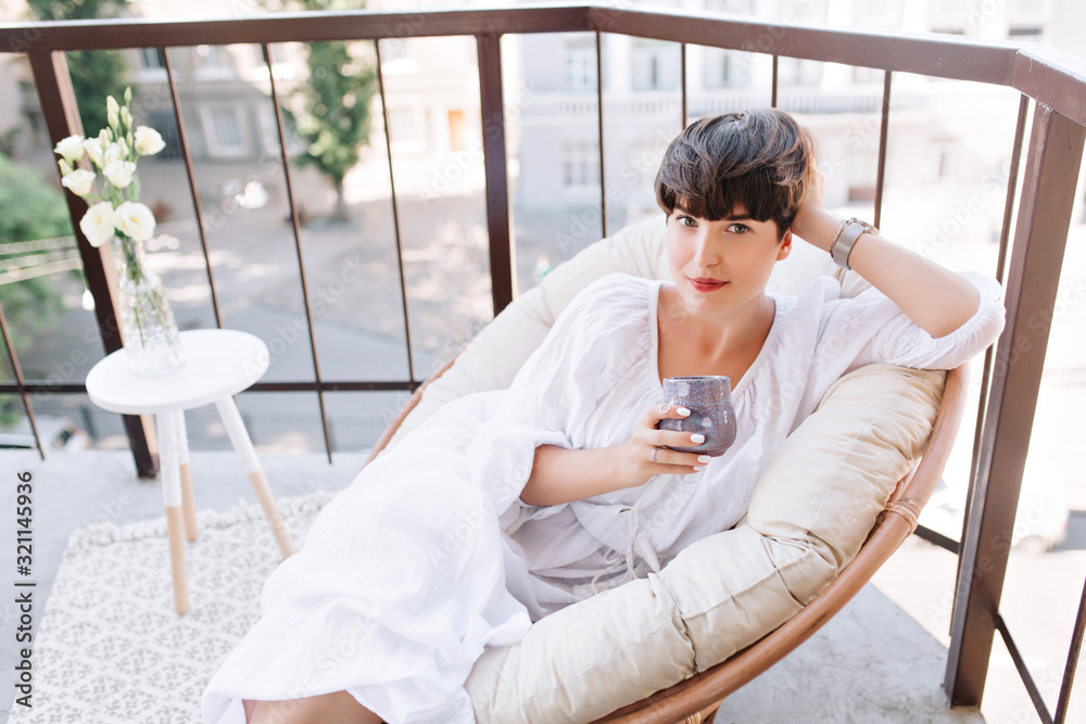 Pretty dark-haired woman in white dress looking with interest holding cup of tea on balcony. Adorable girl with short hair posing while drinking coffee on terrace after breakfast.