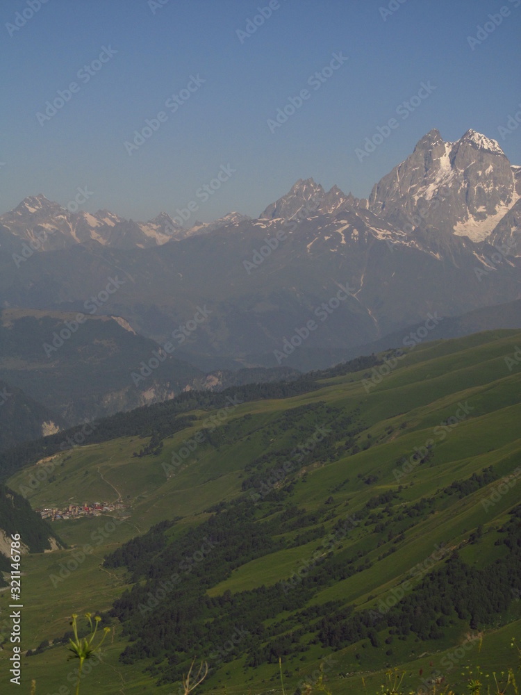 Naklejka premium Mountain landscape of Svaneti on bright summer sunny day. Mountain lake, hills covered green grass on snowy rocky mountains background. Caucasus peaks in Georgia. Amazing view on wild georgian nature