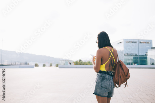 Back view of millennial female tourist with leather backpack standing at urban setting and browsing location app for tracking gps during city sightseeing tour, Generation Z phoning via cellphone