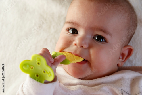 Cute small white skin with brown hair five months baby kid lying on white bed looking up with funny smile on face, holding teether in hand for first teeth, little child with big eyes, innocent girl