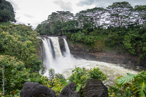 Hilo, Hawaii, USA. - January 14, 2020: Longer shot on White Rainbow Falls on foaming violent Wailuku River surrounded by green trees and plants under white-gray cloudscape.