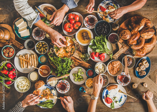 Turkish breakfast table. Flat-lay of peoples hands taking Turkish pastries, vegetables, greens, cheeses, fried eggs, jams and tea in copper pot and tulip glasses over wooden background, top view