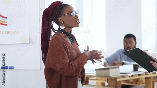 Happy young casual African woman boss in eyeglasses leading discussion, chatting to colleagues at modern light office.