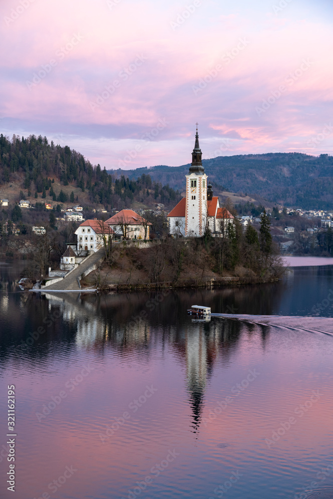 Island Church at Lake Blend in Slovenia during Sunrise. A Pletna boat is transporting tourists.