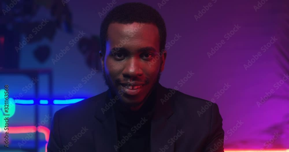 Close up of African American young handsome man smiling to camera in room with neon pink and blue lights. Portrait of black guy in club atmosphere. Male model smile. Close up of African American young handsome man smiling to camera in room with neon pink and blue lights. Portrait of black guy in club atmosphere. Male model smile.