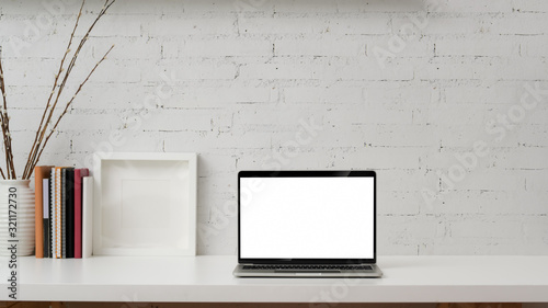 Close up view of blank screen laptop, mock up frame, books and decorations on white table with brick wall background