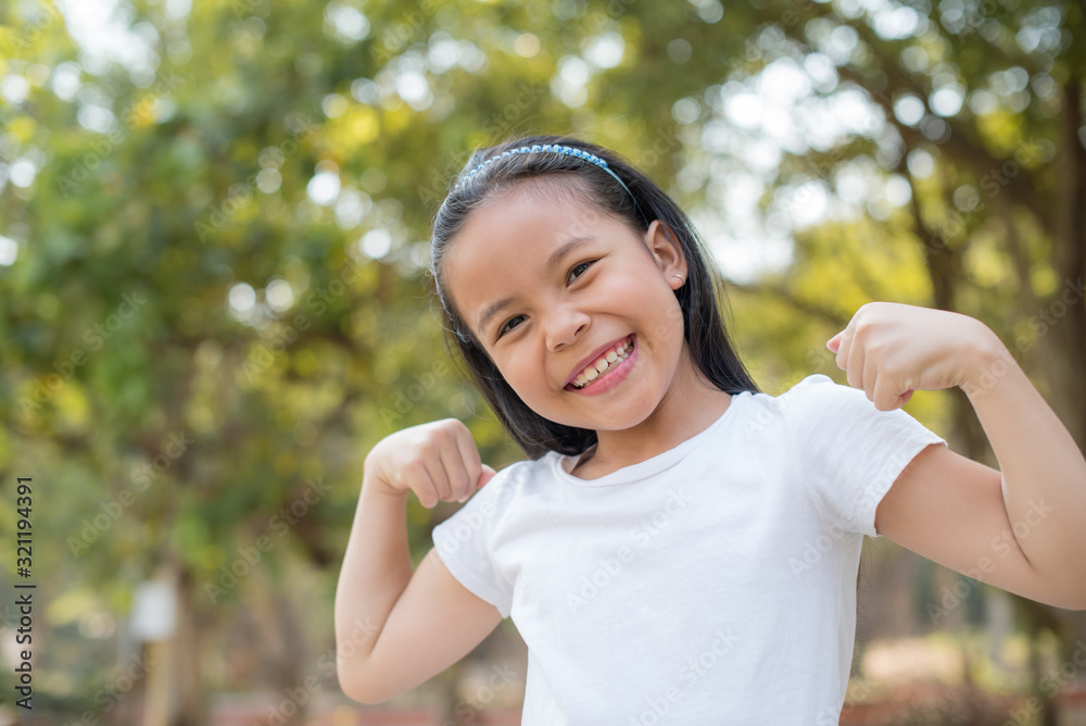 happy Little asian girl child standing showing front teeth with big smile.  showing arms muscles smiling proud. Looking camera showing biceps. fresh healthy green bio background. Fitness concept.