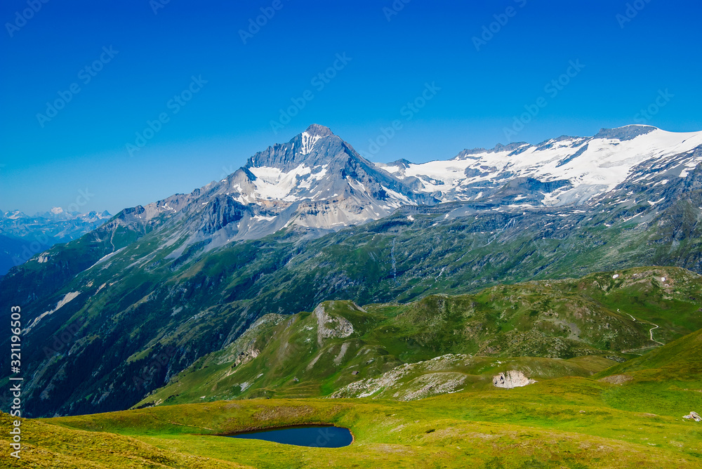 Fototapeta premium Beautiful summer alpine scenery, high mountain with snow on the slopes, green meadow with a small blue lake, hiking trails and clear blue sky in the background, French Alps, Vanoise National Park.
