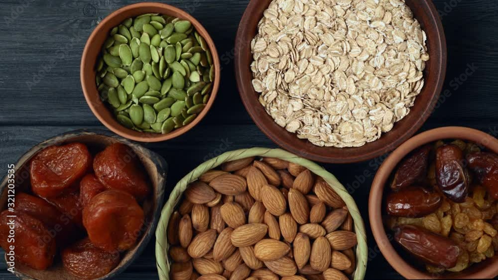 Healthy breakfast. Granola. Oatmeal, barley flakes. Nuts: hazelnuts, almonds, cashews. Dried fruits: raisins, dates, dried apricots. In clay bowls on a black, wooden background. Slow motion.Top view. 