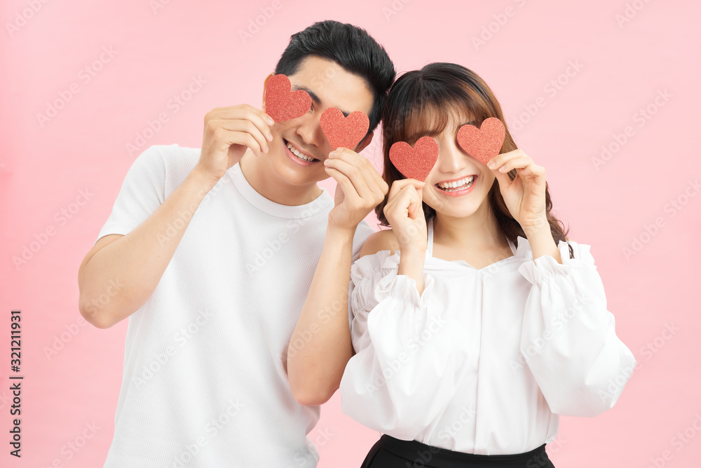 Attractive young couple holding red love hearts over eyes isolated on pink background Attractive young couple holding red love hearts over eyes isolated on pink background