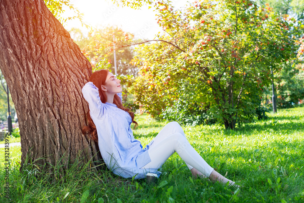 Young woman sitting under tree in summer park - obrazy, fototapety, plakaty 