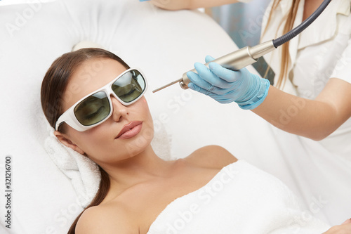 beautiful woman in protection glasses at cosmetology center receiving hair removal for her cheek by professional beautician