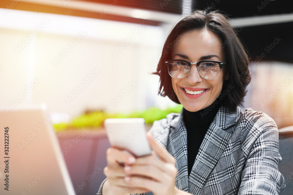 Graceful beauty. Close-up photo of stylish modern business woman with glasses holding smartphone in her hands and smiling while looking in the camera. Graceful beauty. Close-up photo of stylish modern business woman with glasses holding smartphone in her hands and smiling while looking in the camera.