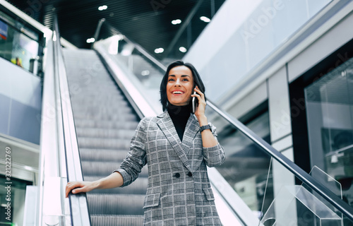 In the mall. Elegant young business woman in smart casual outfit goes down the escalator while talking on the phone.