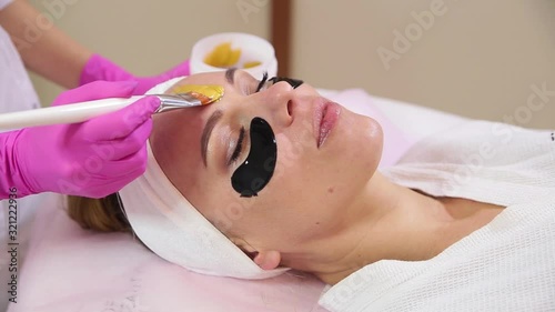 Hand of professional cosmetologist applying gold mask on client's face.
