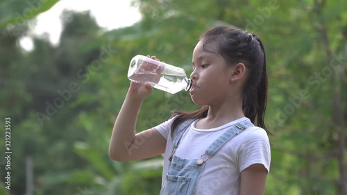 A little girl drinking clean water from a bottle against the background of trees. A happy little girl in a white t-shirt with a long haircut is smiling at the camera on the nature outdoors.