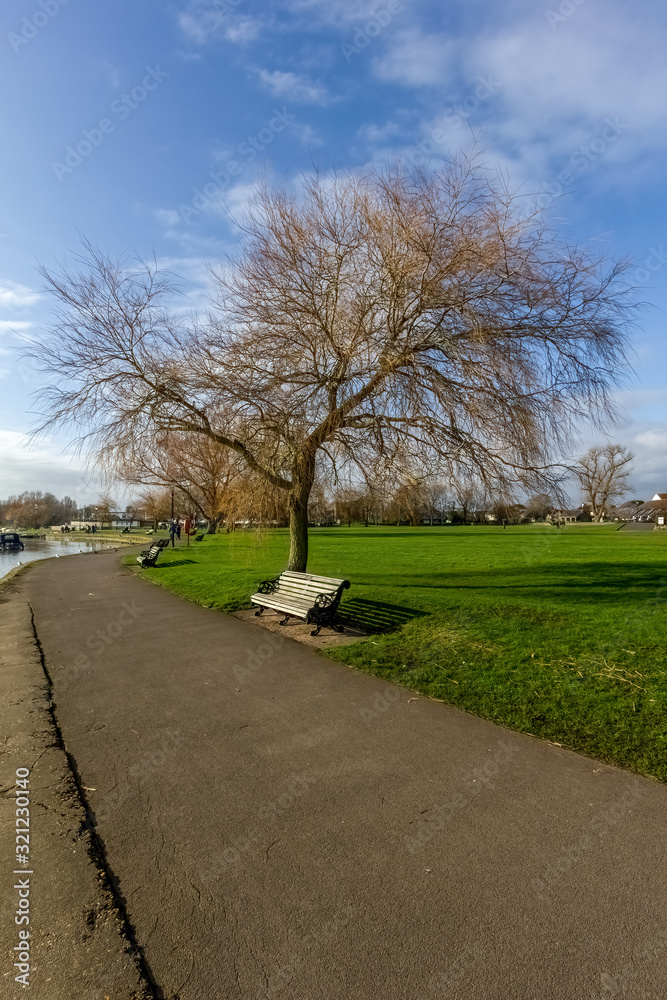 A scenic view of a park path along a water way with trees and bench under a majestic blue sky and white clouds
