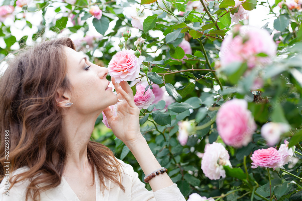 closeup portrait of beautiful young woman brunette with pink rose bush on background. girl stands in profile and reaches for a flower and sniffs it closeup portrait of beautiful young woman brunette with pink rose bush on background. girl stands in profile and reaches for a flower and sniffs it