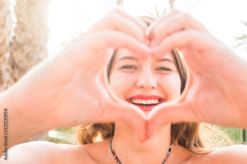 Close up portrait of beautiful.  caucasian young woman doing love hearth sign with hands at the camera - sun bright background and joyful happiness people concept photo