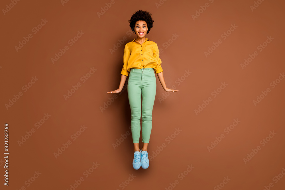 Full body photo of funny dark skin lady jump high up flirty mood toothy smiling wear yellow shirt green pants footwear isolated brown color background Full body photo of funny dark skin lady jump high up flirty mood toothy smiling wear yellow shirt green pants footwear isolated brown color background